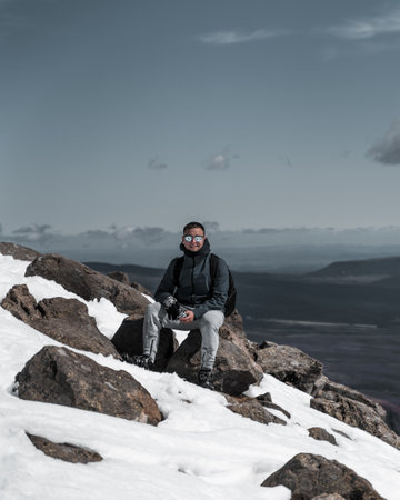 A vertical shot of a young Caucasian cheerful guy sitting on a rock on a snowy mountainの写真素材