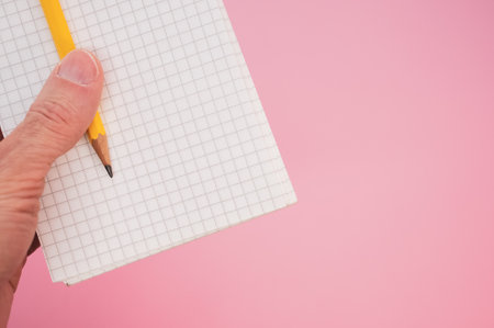 A closeup shot of a hand holding a pencil with a notebook on a pink backgroundの写真素材
