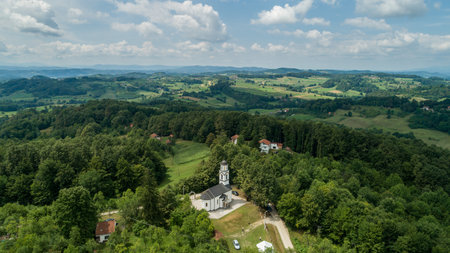 An aerial view of green forests and fields with a church and residential buildings under a clear skyの写真素材