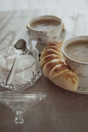 A selective focus shot of two cups of cappuccino with a homemade cheese roll and sugar on a tableの写真素材