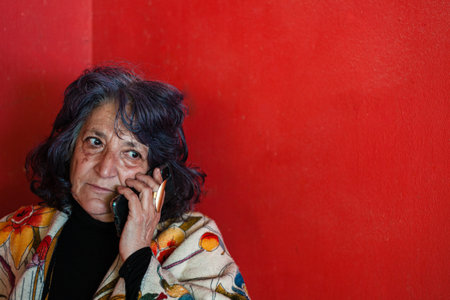 A closeup shot of an elder Hispanic female talking on the phone near a red wall