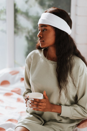 An attractive young African female with long black hair holding a white ceramic mug while sitting on the bedの写真素材