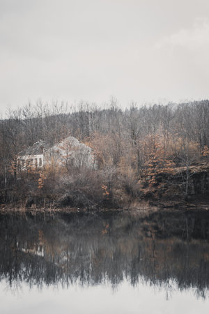 A vertical shot of a beautiful lake surrounded by hill forests and an unfinished houseの写真素材