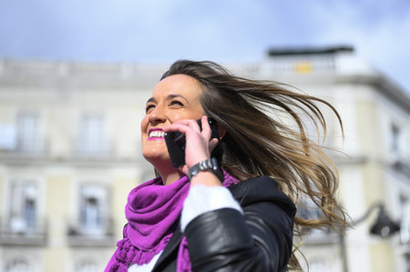 A young Spanish beautiful woman talking on the phone and laughing with a bright purple scarf on a windy dayの写真素材