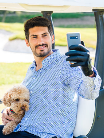 A young Caucasian male in a golf cart taking a selfie with his Goldendoodle puppy on a golf courseの写真素材