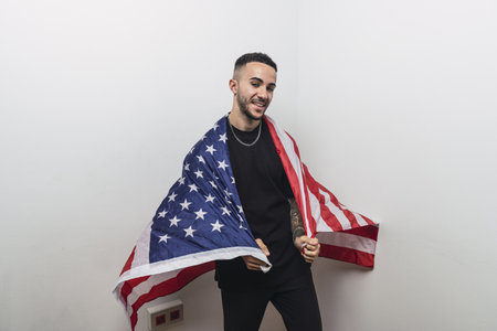 A young proud Hispanic man holding a US flag around is shoulders against a white backgroundの写真素材