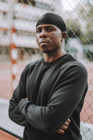 A shallow focus of a Black man in black clothes and do-rag leaning on a cyclone wire fence