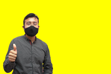 A young attractive man from India in a black facemask showing the thumbs u gesture on a yellow...