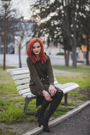 An attractive young Caucasian female with red hair sitting on a bench in a parkの写真素材