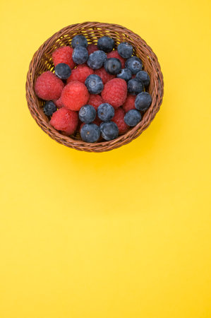 A vertical shot of fresh raspberries and blueberries in a basket isolated on yellow background
