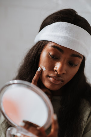A pretty young African female wearing a white headband applying face cream while looking in a hand mirror in the bedroomの写真素材