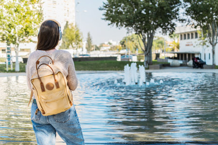 A back view of a young woman with a backpack, coffee, and headphones, walking in a park