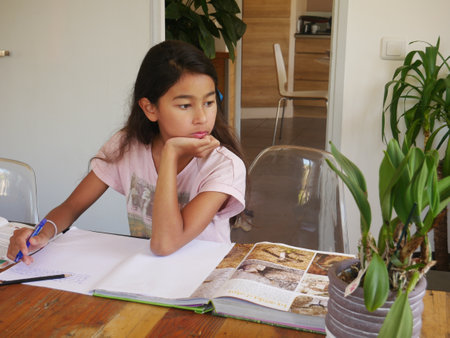 A beautiful girl from France looking right sitting at the table with books and a green plant