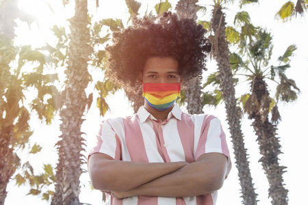 Handsome African young man wearing a pride mask and posing with crossed arms in front of palm trees...
