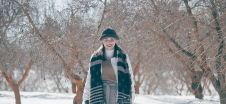 A selective focus shot of a cheerful young lady wearing warm winter clothe in a snowy winter park
