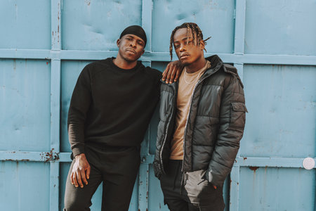 Two young Black Spanish male friends standing in front of a blue metal gate