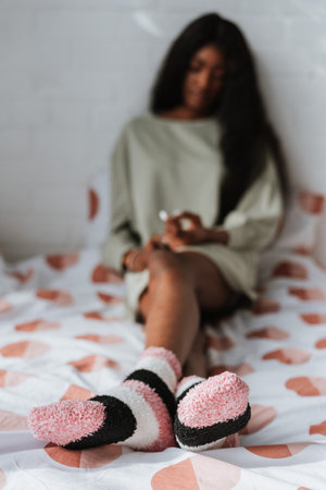 A selective focus shot of a young African female wearing soft striped socks sitting on the bedの写真素材
