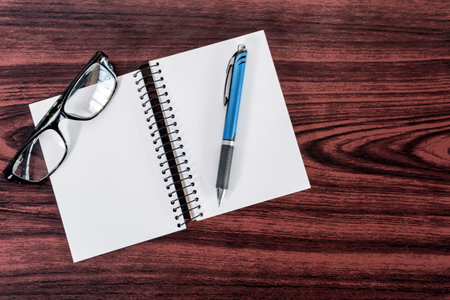 A top view of a notebook, pen, and glasses on a wooden tableの写真素材
