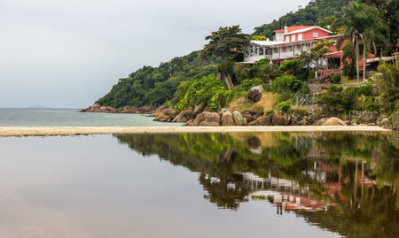A beautiful shot of a seaside house by a beach in Brazil with its reflection visible in the water and tree-covered hills in the backの写真素材