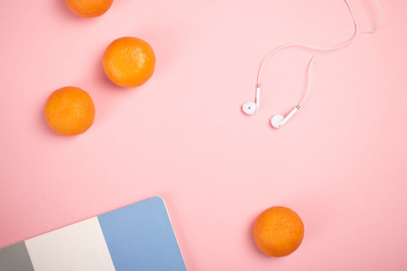 A top view of a notepad, earphones, and tangerines on a pink background