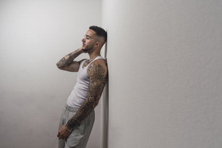 A portrait of a young handsome tattooed Spanish male posing leaning on a white wallの写真素材