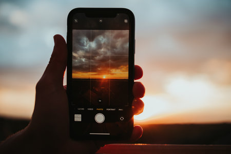 A hand holding a smartphone, taking a picture of lovely golden sunset