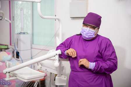 A Mexican female dentist in a purple outfit wearing a mask and goggles leaning on the dental chairの写真素材