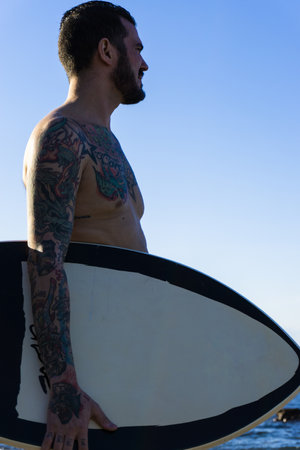 A vertical shot of a Caucasian young surfer male with tattoos near the sea