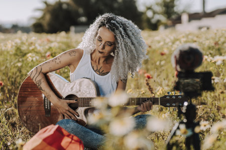 A curly-haired Spanish cute woman playing guitar in a field of daisies while recording a vlogの写真素材