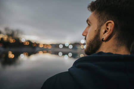 A shallow focus shot of a Caucasian brunette man from the back with a beard and an earring, and a blurred background with a lake and lights.の写真素材
