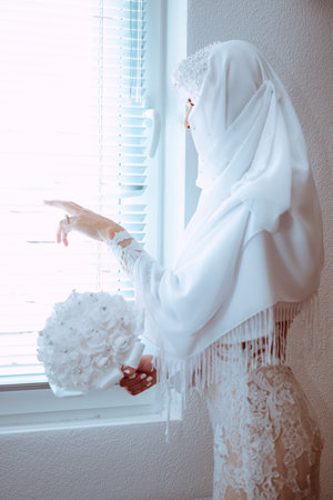 A beautiful bride in a white headscarf looking from the windowの写真素材