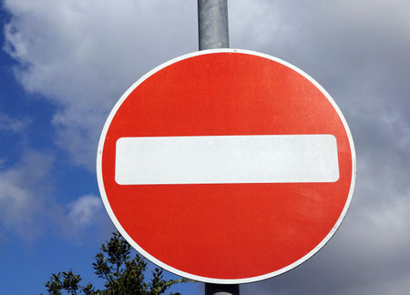 A closeup of a round no entry sign in the street with trees and big clouds in the background
