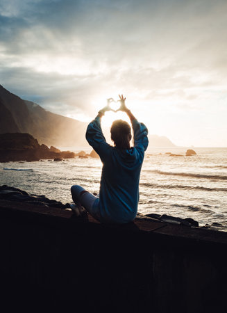 A silhouette of a girl sitting by the sea making a heart with her fingers against an orange sunset skyの写真素材