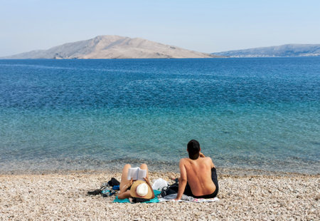 Young couple relaxing on beach and reading a book during summer vacation. Beautiful blue sea and rocky coastline on Pag island in Croatiの写真素材