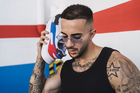 A hot caucasian man with arm tattoos in a black shirt and white pants posing with a sneaker to his earの写真素材