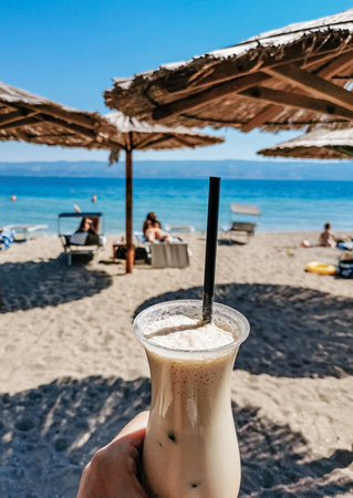 Man holding iced coffee under parasol on sandy beach.の写真素材