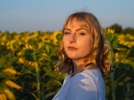 A portrait of a young Caucasian female with a piercing and blonde curly hair in a sunflower field