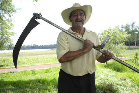 A Hispanic male farmer with an old scythe for cutting weeds on a farmの写真素材