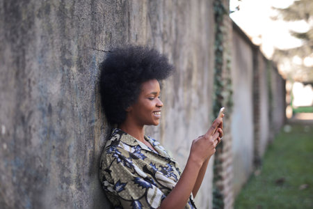 portrait of young black woman leaning against a wall using her smartphoneの写真素材