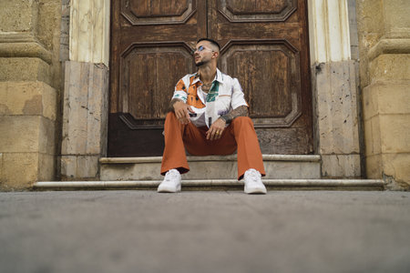 A hot caucasian man with arm tattoos with a patterned shirt and brown pants sitting on the entranceの写真素材