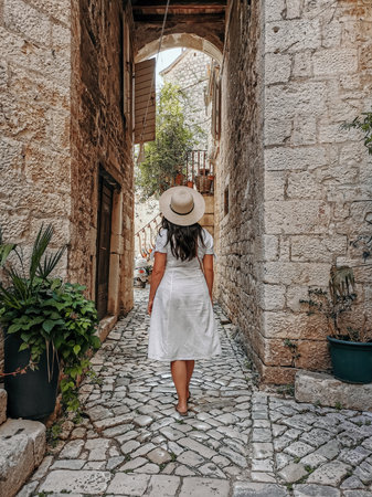 Rear view of young woman wearing white dress and sun hat holding handbag. Lifestyle and fashion full length image of female tourist in old town.の写真素材