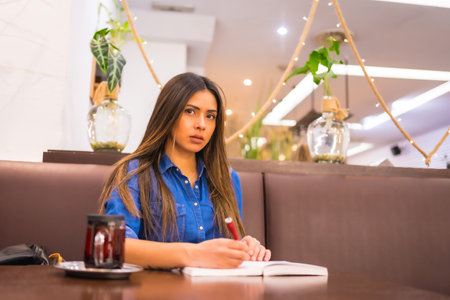 A brunette Caucasian girl in a denim shirt having tea in a coffee shop. Writing in a notebook a work for the universityの写真素材