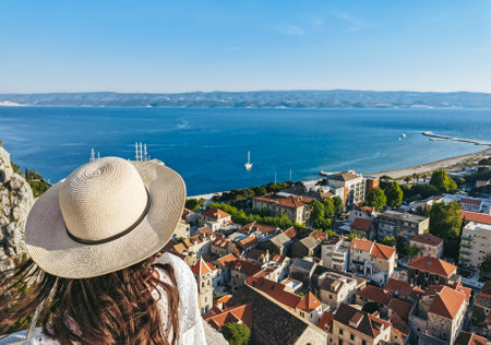 Rear view of young woman wearing stylish white summer dress and sun hat standing on lookout point...