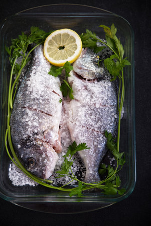 Fish in a glass tray with coarse salt, lemon slices and parsley on a dark background.の写真素材
