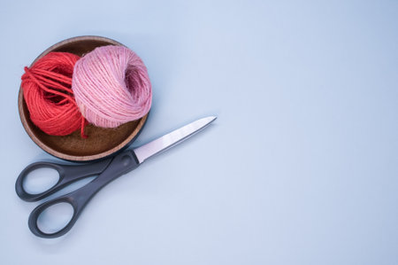A top view of yarns for knitting and scissors in a wooden bowl on a blue background