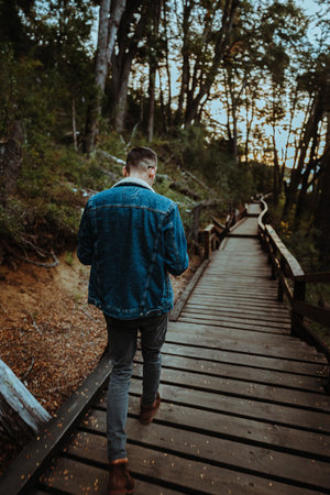 A vertical shot of a stylish young man walking on a long wooden track in the woodsの写真素材