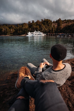 A vertical shot of a couple of Argentine boys sitting at the edge of a lake, admiring the view with...