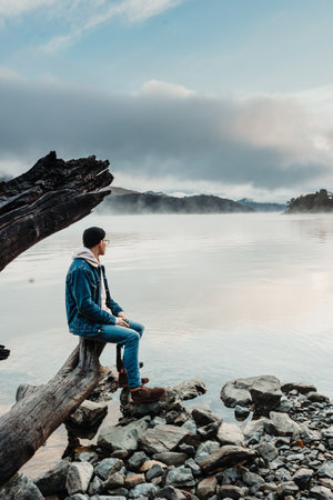 A vertical shot of a stylish Argentinian man in jeans, admiring beautiful view of a calm lake while sitting at the end of a wooden logの写真素材