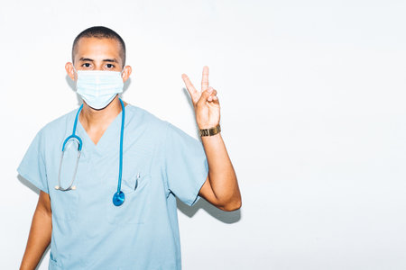 Portrait of young latin american man doctor making peace gesture with his hands. corona virus, hope concept.の写真素材