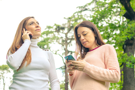 A young woman is talking on her cell phone while her friend is looking at her smart phone.の写真素材